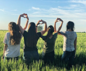 Four girls in a field forming a heart shape with their bodies, surrounded by green grass and bright sunlight.