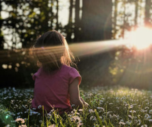 A little girl sits in the grass, illuminated by sunlight shining brightly behind her.
