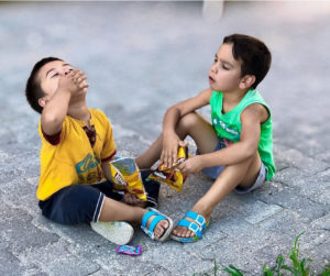 Two boys seated on the ground, sharing and eating candy with joyful expressions.