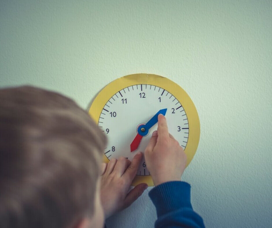 A child holds a clock mounted on a wall, smiling as they point to the time displayed on the clock face