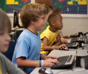 Children engaged with computers in a bright classroom, focused on their screens and collaborating on projects.