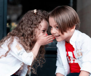 Two children sit on a bench, with one whispering a secret to the other, both smiling and engaged in conversation.