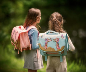 Two girls with backpacks stand beside a pond, looking at the water and enjoying their time outdoors.