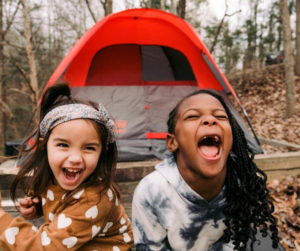 Two young girls laugh joyfully in front of a colorful tent, enjoying a playful moment together outdoors.