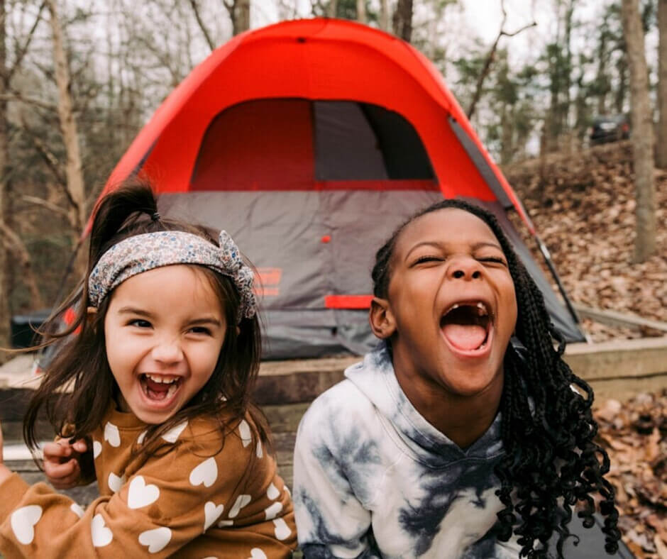 Two young girls laugh joyfully in front of a colorful tent, enjoying a playful moment together outdoors.