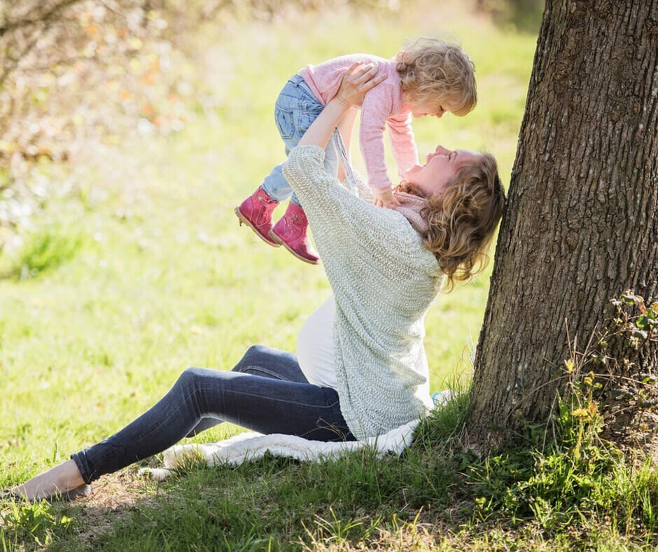 A mother holds her daughter high above her head, both expressing happiness in a playful outdoor setting.