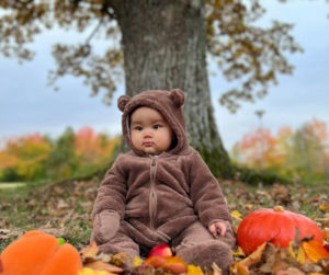 A cheerful baby in a fluffy bear costume sits in a pile of vibrant leaves, surrounded by the beauty of fall.