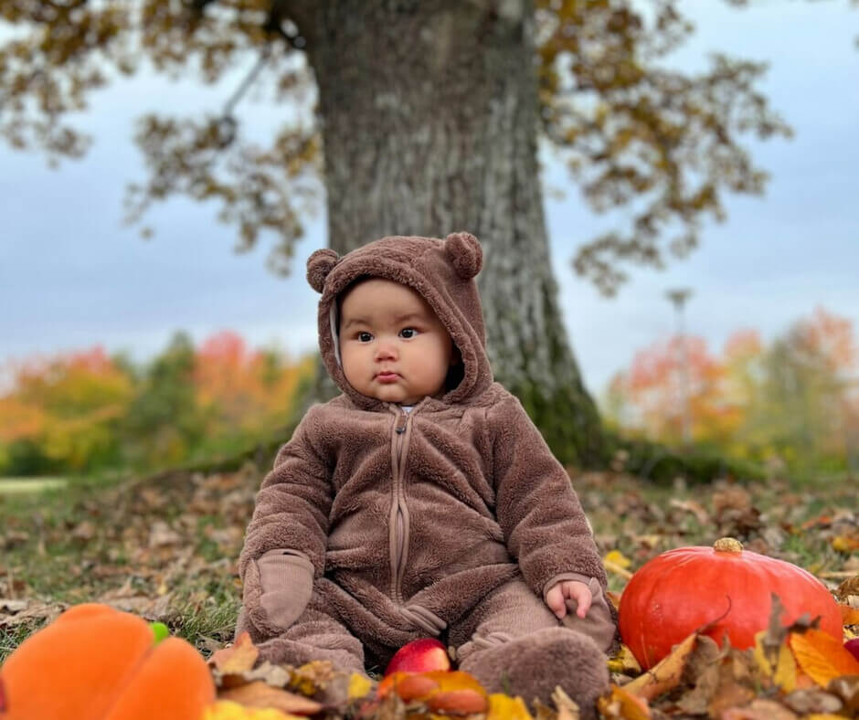 A cheerful baby in a fluffy bear costume sits in a pile of vibrant leaves, surrounded by the beauty of fall.