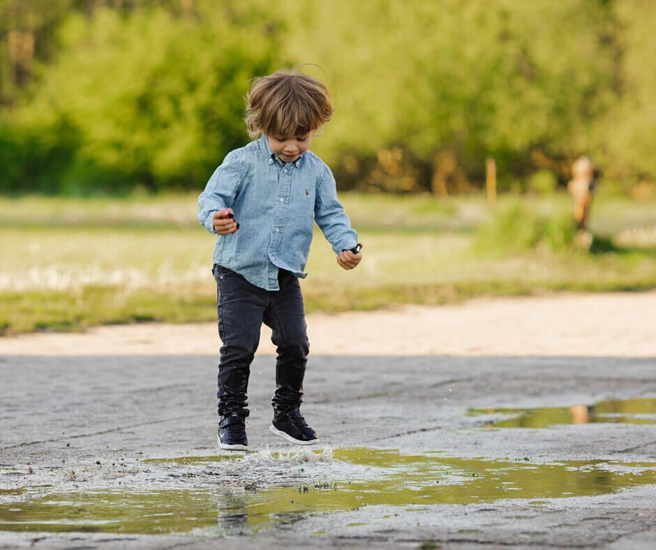 A young boy joyfully jumps into a puddle, splashing water around him on a rainy day.