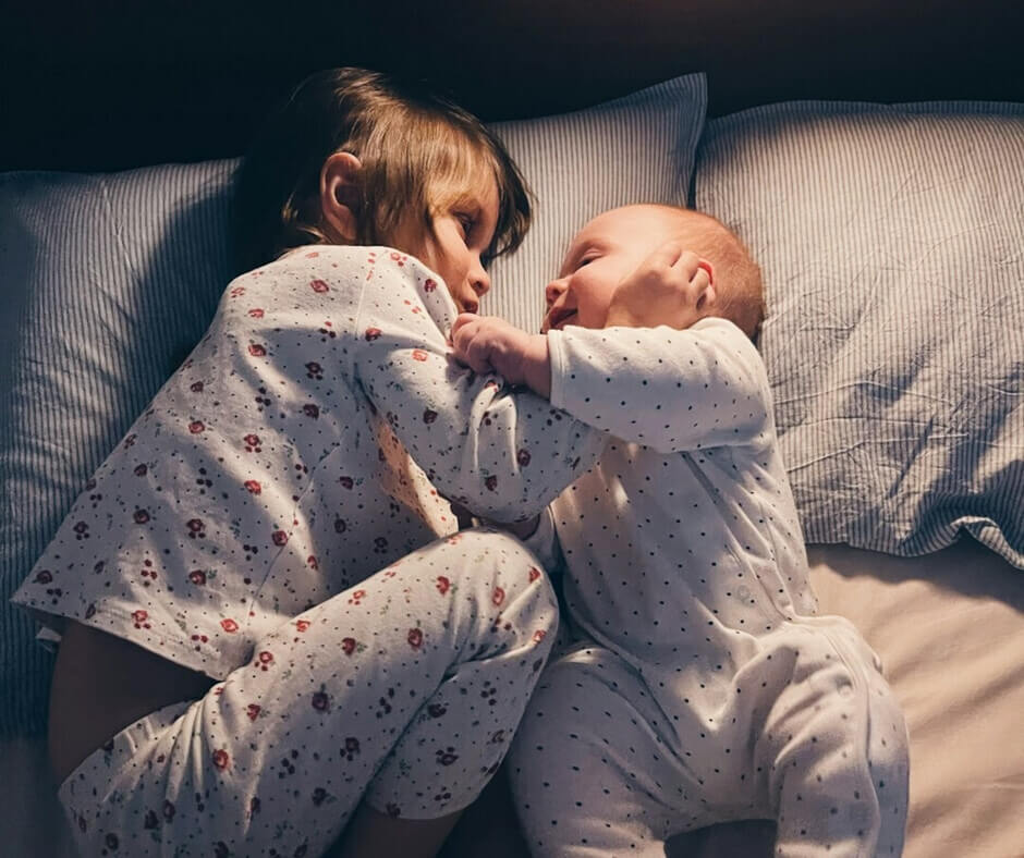 Two kids in matching pajamas relaxing on a bed, surrounded by soft pillows and blankets.