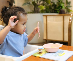 A toddler in a high chair holds a spoon, looking curiously at the food in front of them.