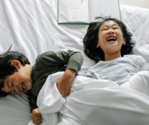 Two children laughing together in bed while reading a book, surrounded by soft pillows and cozy blankets