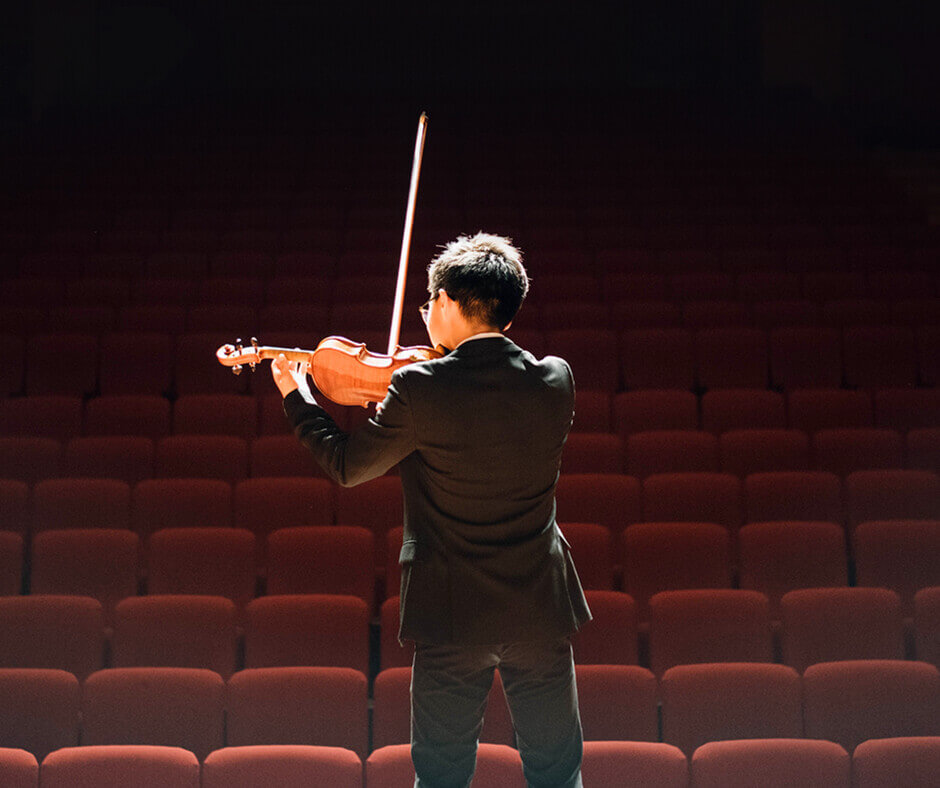 A boy in a suit plays the violin on stage in an auditorium, surrounded by an audience enjoying the performance.