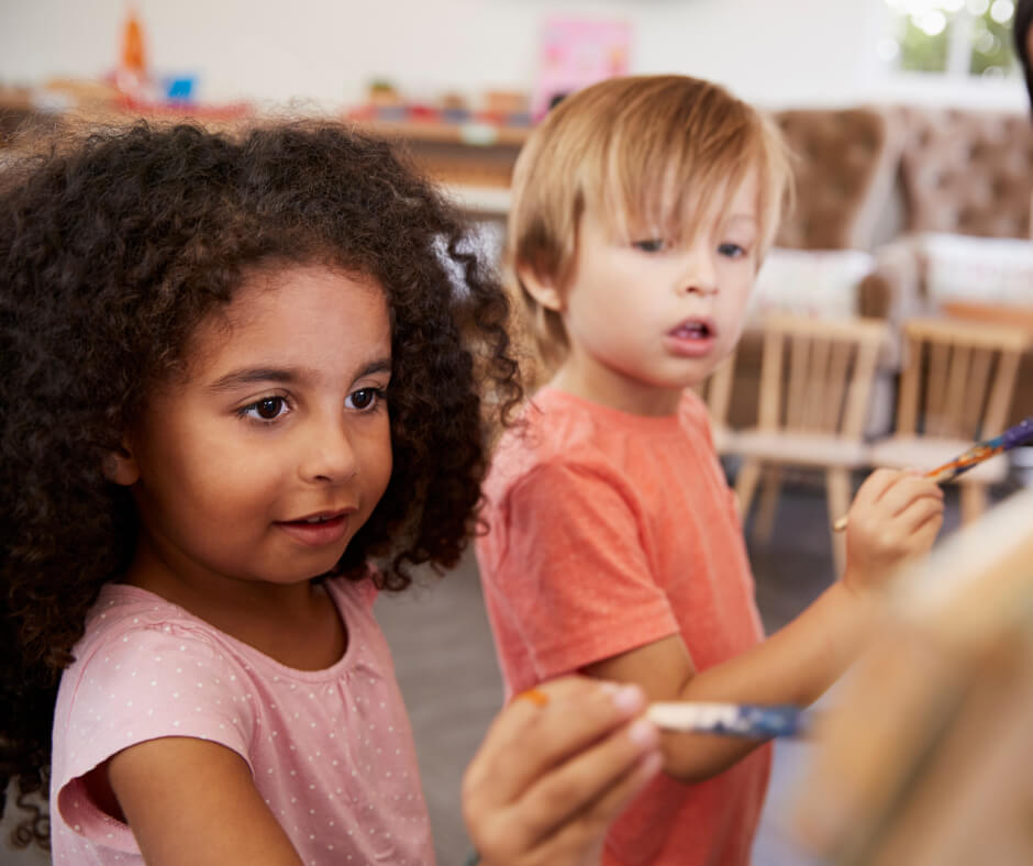 Two children are engaged in painting activities at their classroom desks, surrounded by art supplies and colorful artwork.