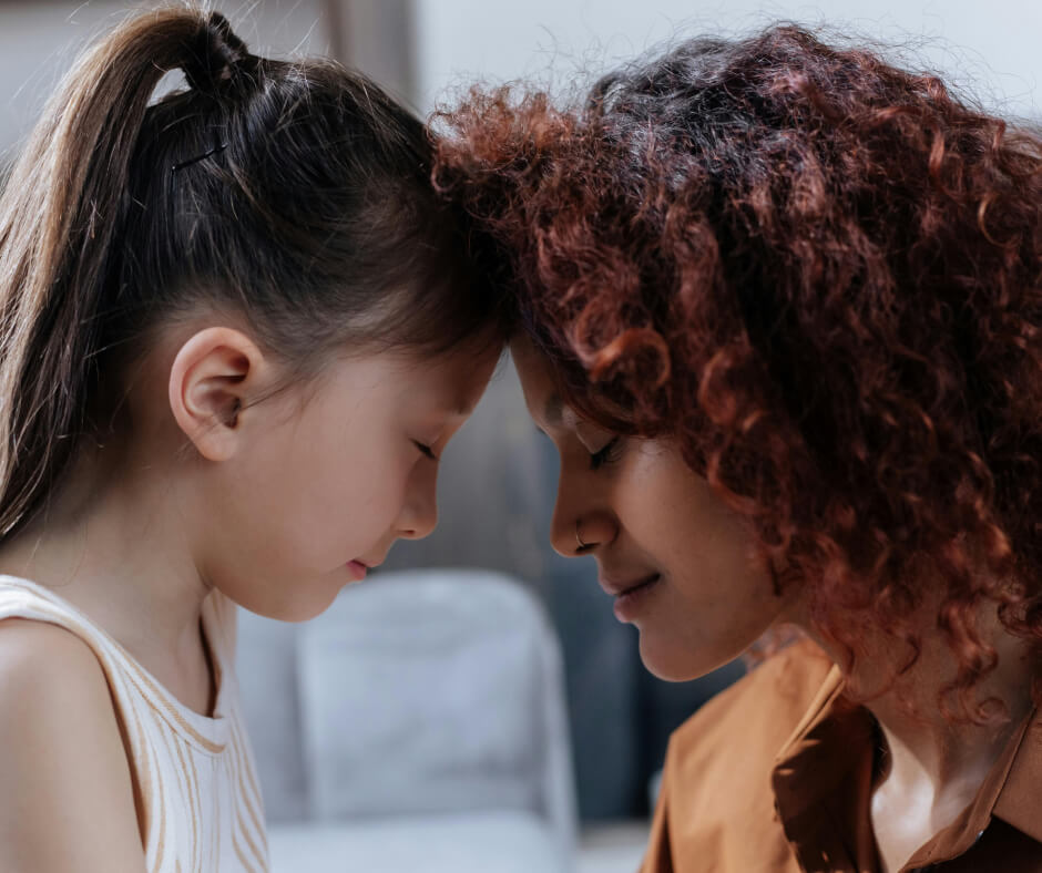A woman and a young girl look at each other, their faces reflecting curiosity and affection in a tender moment.