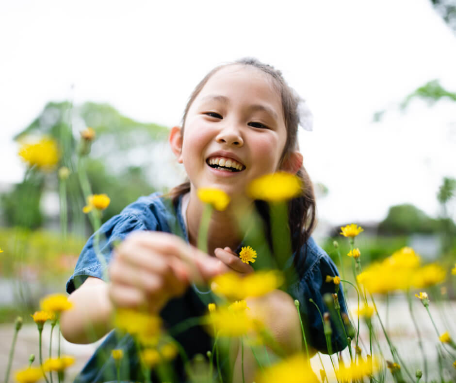 A cheerful young girl admires bright yellow flowers, her smile reflecting joy in a natural setting.