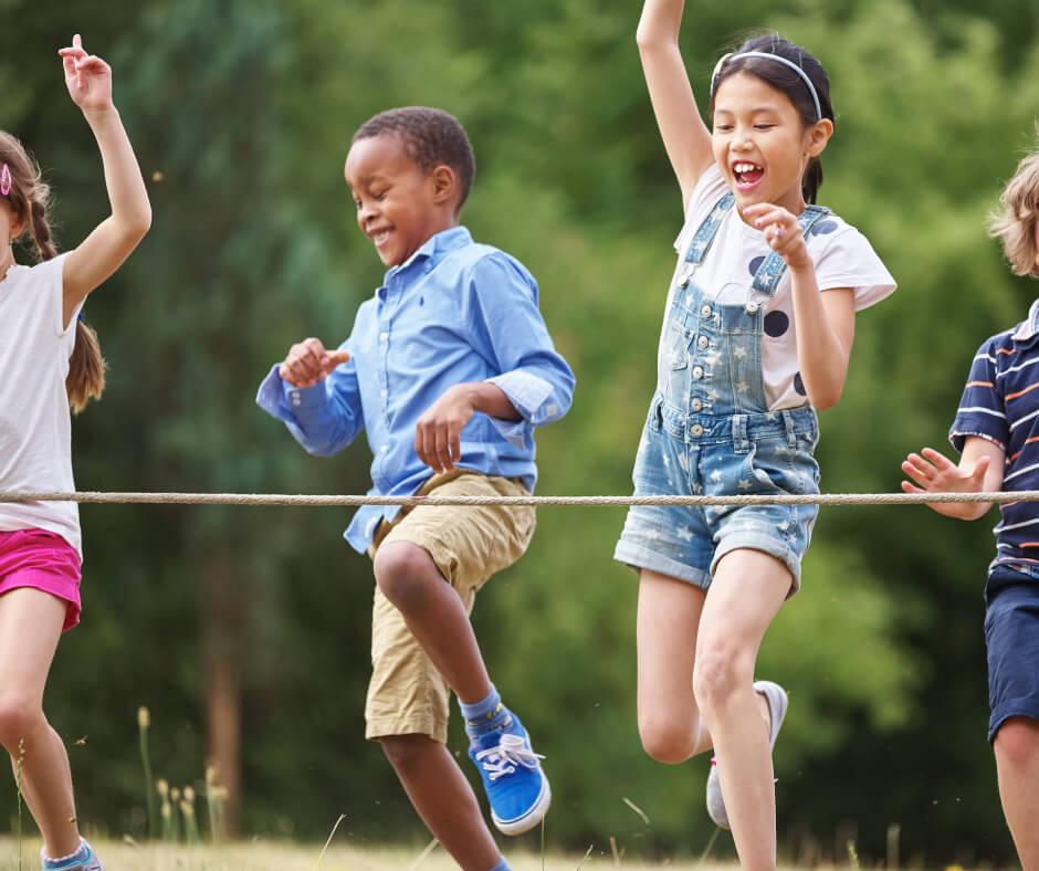 Four children jump over a rope outdoors, smiling and playing together on a sunny day with green trees in the background.