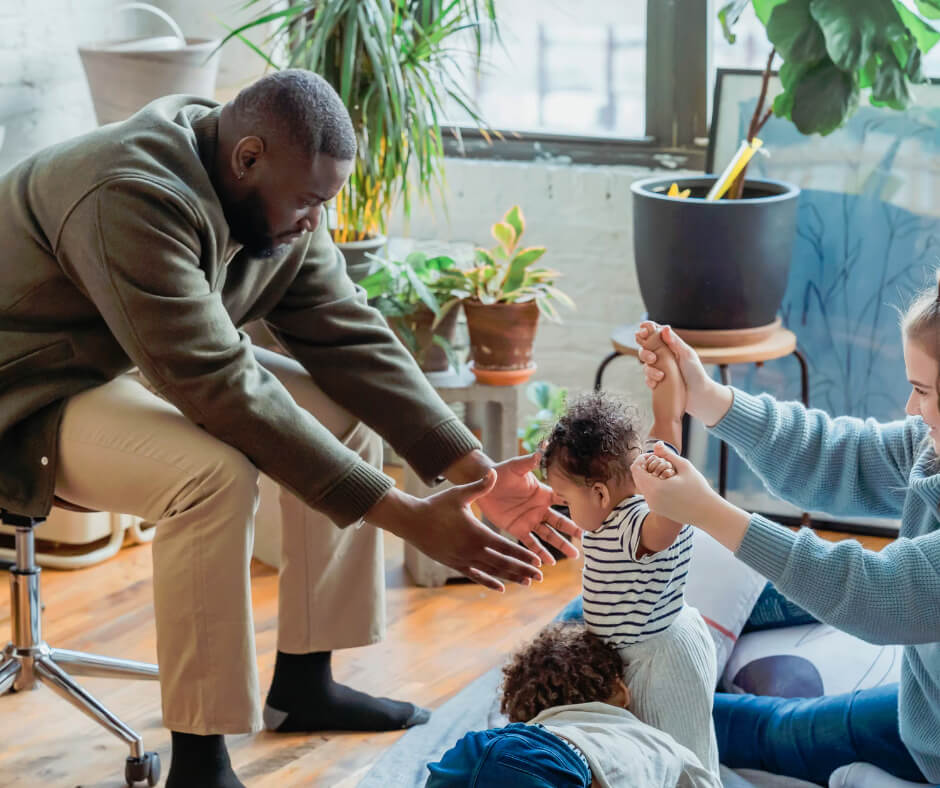 Father encouraging toddler to take first steps while mother watches, demonstrating key developmental milestones in early childhood