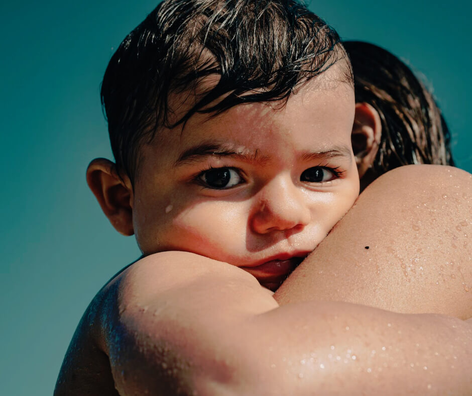 Wet baby with dark eyes resting on parent's shoulder after swimming, showing sun-exposed skin for heat rash vs sunburn comparison