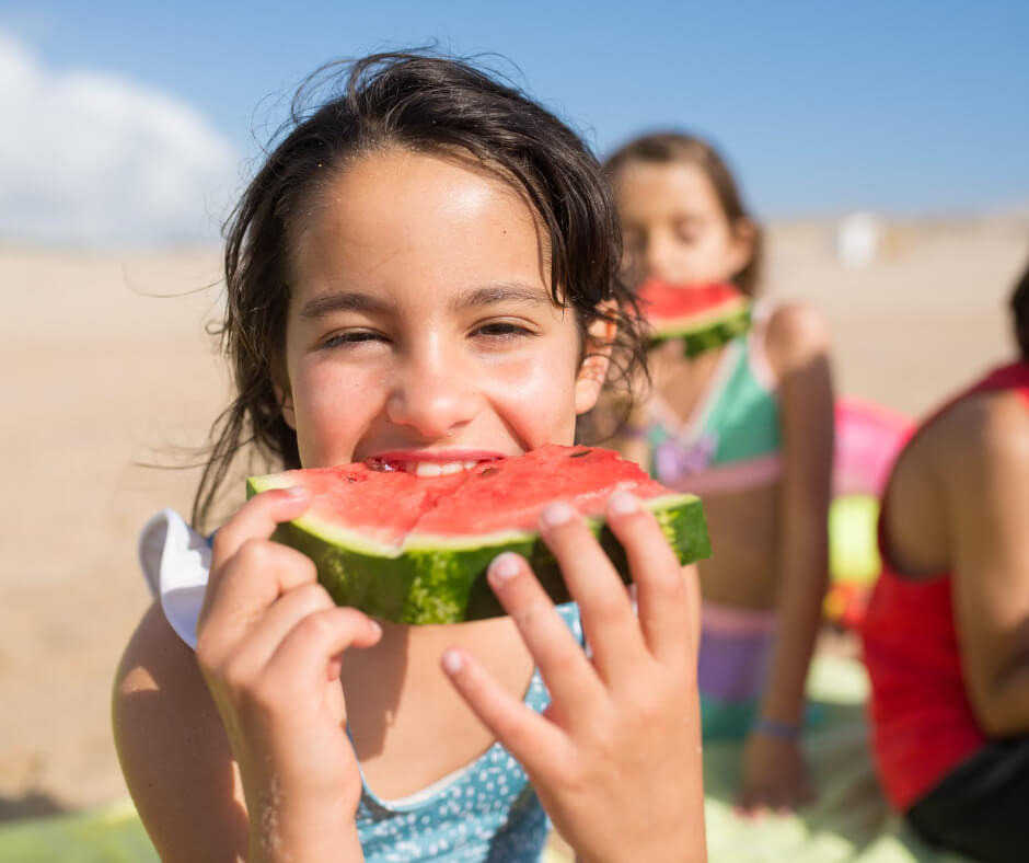 Young girl joyfully eating watermelon slice at beach, healthy hydrating snack for active kids in summer heat