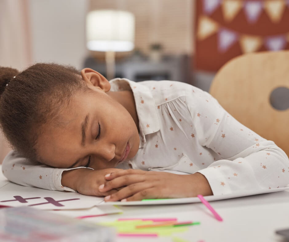 Tired young girl sleeping at desk with schoolwork, illustrating sugar crash fatigue in children during summer