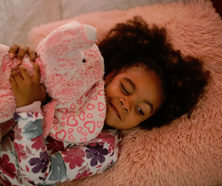 Young girl in floral pajamas hugging pink stuffed animal on fluffy bed, illustrating children's sleep and bedwetting comfort