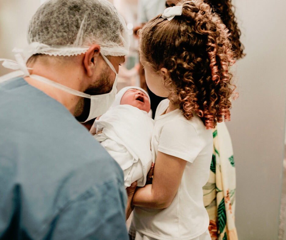 Young girl holds newborn sibling in hospital while doctor looks on, illustrating sibling adjustment after new baby arrives